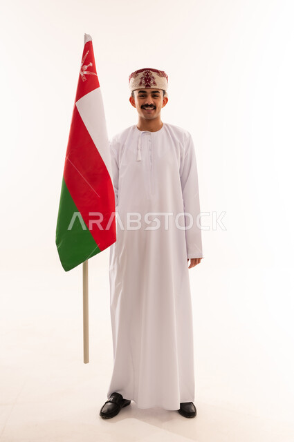 المشاركة بالاعياد والمناسبات الوطنية، الاحتفال بذكرى اليوم الوطني العماني، الاعتزاز والفخر بالرموز والراية العمانية، بورتريه لشاب عربي خليجي عماني يرتدي الدشداشة والكمه يحمل بيده علم سلطنة عمان، مفهوم حب الوطن والإنتماء إليه، الجسم بالطول الكامل، خلفية بيضاء