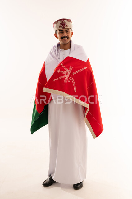 الفخر والاعتزاز بالانتماء للهوية، المشاركة بالاعياد والمناسبات الوطنية، الاحتفال بذكرى اليوم الوطني العماني، الاعتزاز والفخر بالرموز والراية العمانية، بورتريه لشاب عربي خليجي عماني يرتدي الدشداشة والكمه، مفهوم حب الوطن، الجسم بالطول الكامل، خلفية بيضاء