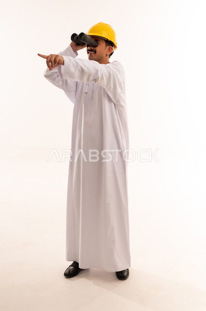 Seeing distant objects through a telescope, future planning for goals and ambitions, love of exploration and adventure, portrait of a university engineering student wearing the Saudi Arabian Gulf dress and helmet holding binoculars and pointing with his index finger to a place, full body length, white background