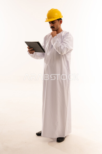 Facial and hand gestures indicating thinking and confusion, hobby of reading and exploration, portrait of a university engineering student wearing Saudi Arabian Gulf clothing and helmet holding a portable tablet in his hand, full body, white background