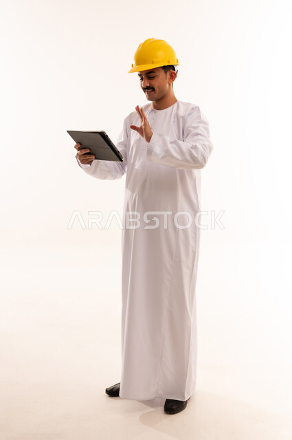 الاستفادة من الأجهزة والتكنولوجيا الحديثة في الاطمئنان على الأقارب، قضاء وقت الفراغ بالتعلم والثقافة، بورتريه لطالب جامعي هندسي يرتدي الزي العربي الخليجي السعودي و الخوذة يحمل بيده جهاز التابلت المحمول، الجسم بالطول الكامل، خلفية بيضاء