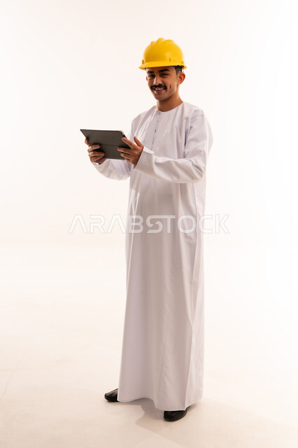 تصفح مواقع التواصل الاجتماعي، قضاء وقت الفراغ بالتعلم والثقافة، هواية المطالعة والاستكشاف ، بورتريه لطالب جامعي هندسي يرتدي الزي العربي الخليجي السعودي و الخوذة يحمل بيده جهاز التابلت المحمول ويقوم بالعمل عليه ،الجسم بالطول الكامل، خلفية بيضاء