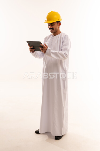 مفهوم التثقيف والبحث العلمي، قضاء وقت الفراغ بالتعلم والثقافة، هواية المطالعة والاستكشاف ، بورتريه لطالب جامعي هندسي يرتدي الزي العربي الخليجي السعودي و الخوذة يحمل بيده جهاز التابلت المحمول ويقوم بالعمل عليه ،الجسم بالطول الكامل، خلفية بيضاء