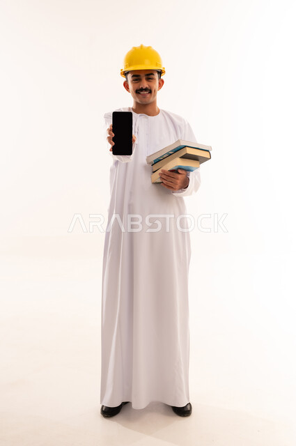 تعابير السعادة والفرح، استخدام الأجهزة والتقنيات الحديثة، بورتريه لطالب هندسي عربي خليجي عماني يرتدي الدشداشة و الكمه يمسك بيده الهاتف المحمول يقوم بعرض شاشة سوداء فارغة بإيماءات السرور، صورة الجسم بالطول الكامل, خلفية بيضاء