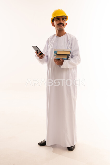 التواصل مع الأهل والأصدقاء وسماع أخبارهم، الاستفادة من الأجهزة والتكنولوجيا الحديثة في الاطمئنان على الأقارب، بورتريه لطالب جامعي هندسي يرتدي الزي العربي الخليجي السعودي و الخوذة يمسك الهاتف المحمول و يقوم بالنظر الى الاعلى، الجسم بالطول الكامل، خلفية بيضاء