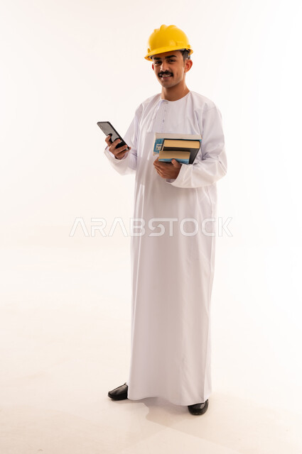 الاستفادة من الأجهزة والتكنولوجيا الحديثة في الاطمئنان على الأقارب، التواصل مع الأهل والأصدقاء وسماع أخبارهم، بورتريه لطالب جامعي هندسي يرتدي الزي العربي الخليجي السعودي و الخوذة يمسك الهاتف المحمول بإيماءات السعادة، الجسم بالطول الكامل، خلفية بيضاء
