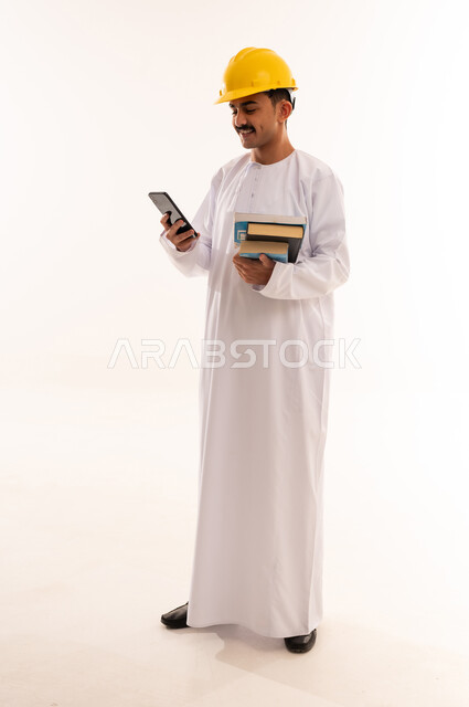 الإستعداد للمحاضرات الجامعية الإلكترونية ، بورتريه لطالب جامعي هندسي يرتدي الزي العربي الخليجي السعودي و الخوذة  يقوم بإجراء مكالمة فيديو من خلال جهاز الموبايل بإيماءات وجه تدل على السعادة، الجسم بالطول الكامل، خلفية بيضاء