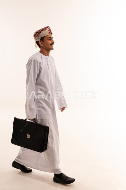 الاهتمام بالأناقة والمظهر الخارجي، الذهاب إلى دوام العمل بنشاط, الشعور بالسرور لتحقيق أهداف الشركة, بورتريه من الجانب لرجل أعمال عربي خليجي عماني مبتسم يرتدي الكمه والثوب التقليدي يحمل حقيبة يد، صورة الجسم بالكامل, خلفية بيضاء