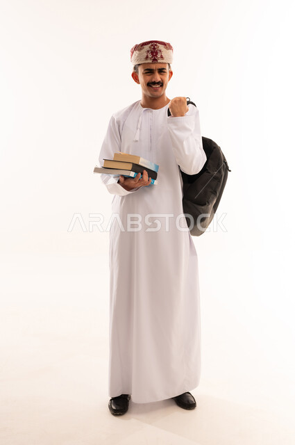 التعليم الجامعي في عمان ، إيماءات وجه تدل على الفرح والنجاح ، تدوين الملاحظات وكتابة تقارير وأبحاث علمية ، بورتريه لطالب جامعي عربي عماني يرتدي الزي التقليدي و يحمل بيده كتب ، مراجعة الدروس اليومية ،الجسم بالطول الكامل، خلفية بيضاء