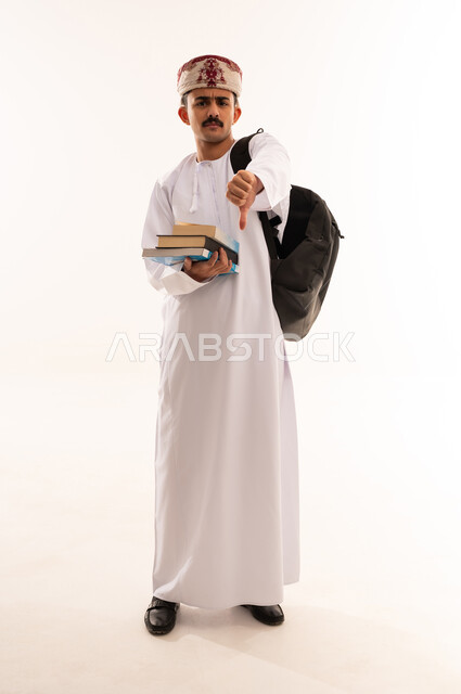 مفهوم الرفض وعدم القبول، بورتريه لطالب عربي خليجي عماني يرتدي الدشداشة و الكمه و الحقيبة يشير بإصبع الابهام للاسفل بإيماءات عدم الاعجاب والتقييم السلبي، الشعور بالانزعاج من شيء ما، صورة الجسم بالطول الكامل, خلفية بيضاء