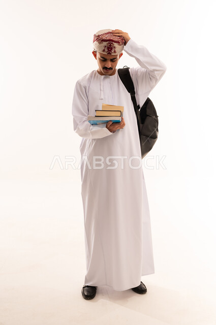 إيماءات وجه ويدين تدل على التذمر والشعور بالإحباط والحزن من الضغط الدراسي ، تدوين الملاحظات وكتابة تقارير وأبحاث علمية ، التعليم الجامعي في عمان ، بورتريه لطالب جامعي عربي يرتدي الزي الخليجي العماني يحمل بيده كتب ،الجسم بالطول الكامل، خلفية بيضاء