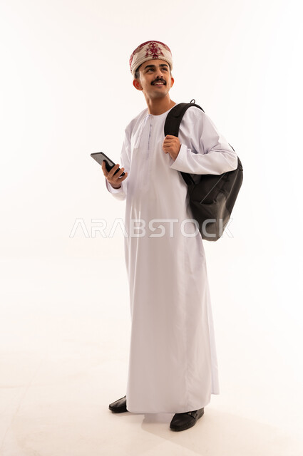 الاستفادة من الأجهزة والتكنولوجيا الحديثة في الاطمئنان على الأقارب، التواصل مع الأهل والأصدقاء وسماع أخبارهم، بورتريه لرجل عربي خليجي عماني يرتدي الدشداشة والكمه ويمسك الهاتف المحمول ينظر الى الاعلى بإيماءات السعادة، الجسم بالطول الكامل، خلفية بيضاء