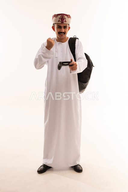 مفهوم الفوز والخسارة ، بورتريه لشاب عربي عماني خليجي يرتدي الزي التقليدي يمسك بيده عصا التحكم و يلعب بألعاب الفيديو ، إيماءات وجه ويدين تدل على الإندماج والتفاعل مع اللعبة ، ألعاب البلايستيشن الترفيهية والممتعة ، قضاء وقت ممتع ، الجسم بالطول الكامل، خلفية بيضاء