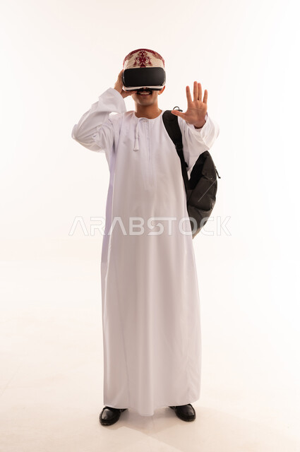 التفاعل والاندماج ومحاكاة العالم الخيالي, بورتريه لشاب عربي خليجي عماني يرتدي الدشداشه والكمه ونظارة الواقع الافتراضي يضع حقيبة الظهر المدرسية, مشاهدة الافلام ثلاثية الابعاد, قضاء أوقات الفراغ بالأنشطة الالكترونية المسلية, الجسم بالطول الكامل, خلفية بيضاء