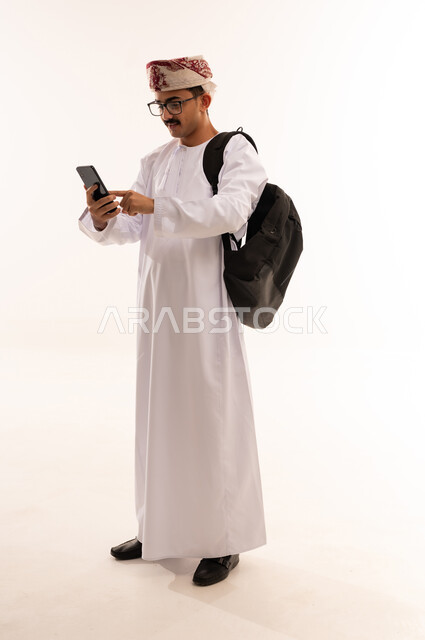 إيماءات وجه تدل على  التركيز، متابعة الدروس اليومية، الإستعداد للمحاضرات الجامعية الإلكترونية ، بورتريه لطالب جامعي يرتدي الزي العربي الخليجي العماني يمسك بجهاز الهاتف المحمول ، التعليم في عمان ، الجسم بالطول الكامل، خلفية بيضاء