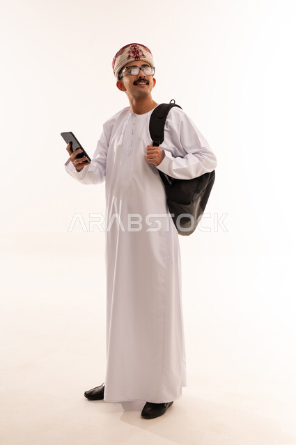 الاستفادة من الأجهزة والتكنولوجيا الحديثة في الاطمئنان على الأقارب، التواصل مع الأهل والأصدقاء وسماع أخبارهم، بورتريه لرجل عربي خليجي عماني يرتدي الدشداشة والكمه ويمسك الهاتف المحمول ينظر الى الاعلى بإيماءات السعادة، الجسم بالطول الكامل، خلفية بيضاء