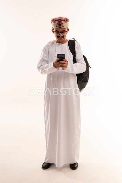 الإستعداد للمحاضرات الجامعية الإلكترونية ، بورتريه لطالب جامعي يرتدي الزي العربي الخليجي العماني يقوم بإجراء مكالمة فيديو من خلال جهاز الموبايل بإيماءات وجه تدل على السعادة ، متابعة الدروس اليومية ، التعليم في عمان ، الجسم بالطول الكامل، خلفية بيضاء