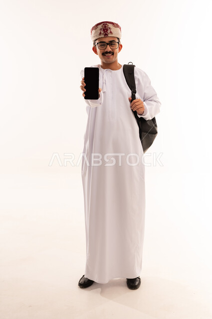 تعابير السعادة والفرح، استخدام الأجهزة والتقنيات الحديثة، بورتريه لشاب عربي خليجي عماني يرتدي الدشداشة و الكمه يمسك بيده الهاتف المحمول يقوم بعرض شاشة سوداء فارغة بإيماءات السرور، صورة الجسم بالطول الكامل, خلفية بيضاء