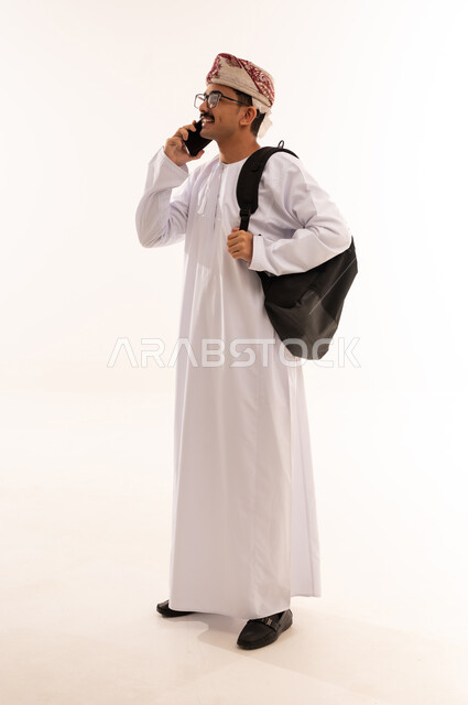 الدردشة والتواصل مع الاصدقاء، استخدام الأجهزة والتقنيات الحديثة، بورتريه لشاب عربي خليجي عماني يرتدي الدشداشة و الكمه يحمل الكتب ويمسك بيده الهاتف المحمول يقوم بإجراء المكالمات الهاتفية، صورة الجسم بالطول الكامل، خلفية بيضاء
