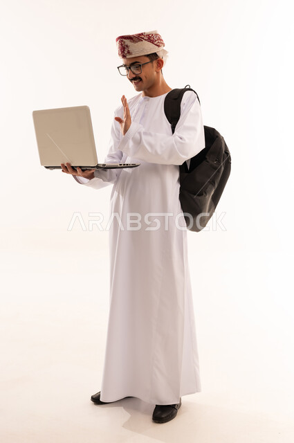 إيماءات تدل على السعادة ، إستخدام أجهزه تقنية حديثة ، بورتريه لطالب جامعي عربي يرتدي الزي الخليجي العماني و الحقيبة يقوم بإجراء مكالمة فيديو من خلال كمبيوتر محمول ، الإستعداد للمحاضرات الجامعية الالكترونية ، التعلم عن بعد ، الجسم بالطول الكامل، خلفية بيضاء