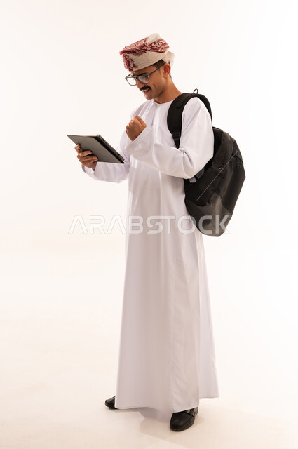 النجاح في الاختبار الجامعي الالكتروني، بورتريه لطالب جامعي عربي خليجي عماني يرتدي الزي التقليدي يحمل بيده جهاز التابلت  ، ايماءات الوجه واليد تدل على الفوز والنجاح ، حل الواجبات الجامعية الالكترونية ، الجسم بالطول الكامل، خلفيه بيضاء