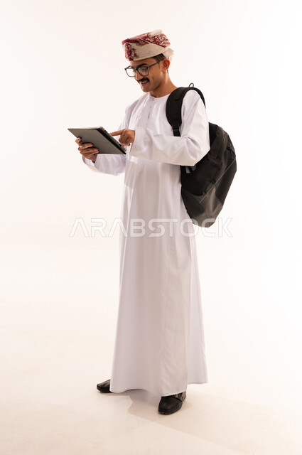 Hobbies of reading, exploration, and spending free time learning and culture, the concept of education and scientific research, portrait of a smiling Omani Gulf Arab man wearing a dishdasha, a sleeve, and a leather bag, holding a portable tablet in his hand and working on it, full-length body image, white background