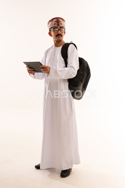 دمج التكنولوجيا في المجال التعليمي،مفهوم التثقيف والبحث العلمي، بورتريه لرجل عربي خليجي عماني يرتدي الدشداشه والكمه وحقيبة جلدية يحمل بيده جهاز التابلت المحمول و يقوم بالنظر الى شيء ما، صورة الجسم بالطول الكامل, خلفية بيضاء