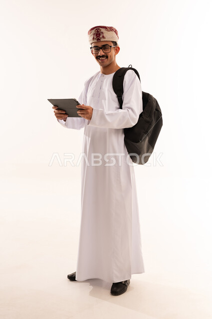 The concept of education and scientific research, the hobby of reading, exploration and spending free time learning and culture, a portrait of a smiling Omani Gulf Arab man wearing a dishdasha, a cufflink and a leather bag, holding a portable tablet in his hand and working on it, a full-length body image, white background