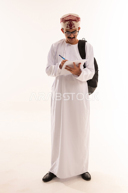 الكتابة وتحديد شيء ما, تدوين وتسجيل الملاحظات، بورتريه لشاب عربي خليجي عماني يرتدي الدشداشة والكمه و النظارة الطبية يمسك بيده قلم يكتب شيء ما, مفهوم التعليم بالمناهج المقررة، الجسم بالطول الكامل، خلفية بيضاء