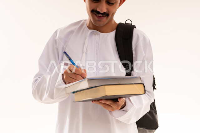مفهوم المذاكرة و الاجتهاد، الاستعداد للعام الجامعي الجديد, الجامعات والكليات الأكاديمية في سلطنة عمان, بورتريه مقرب  لطالب عربي خليجي عماني يرتدي الدشداشة والكمة يحمل حقيبة على ظهره يمسك بيده ورقة وقلم يقوم بمتابعة الدروس اليومية, الجسم بالطول الكامل, خلفية بيضاء