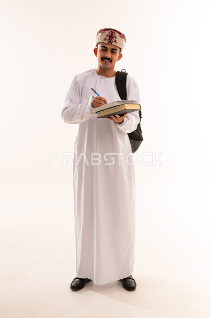 متابعة الدروس اليومية، مفهوم المذاكرة و الاجتهاد، الاستعداد للعام الجامعي الجديد, الجامعات والكليات الأكاديمية في سلطنة عمان, بورتريه مقرب  لطالب عربي خليجي عماني يرتدي الدشداشة والكمة يحمل حقيبة على ظهره يمسك بيده ورقة وقلم، الجسم بالطول الكامل, خلفية بيضاء