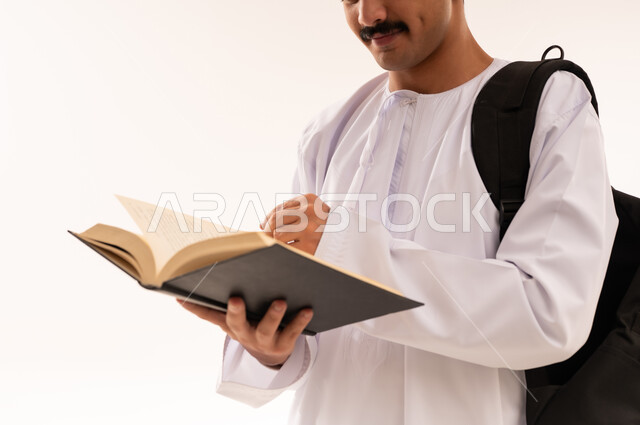 مفهوم الاجتهاد والتفوق، الاهتمام بتصفح الكتب المفيدة، التعليم الجامعي في سلطنة عمان, مراجعة الدروس اليومية, بورتريه مقرب لطالب جامعي عربي خليجي عماني يرتدي الدشداشة والكمة يحمل حقيبة على ظهره وبيده كتاب ويقوم بدراسة المحاضرات,الجسم بالطول الكامل، خلفية بيضاء