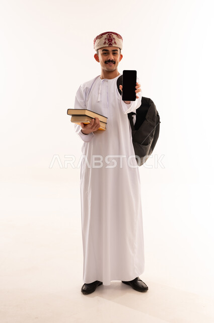 استخدام الأجهزة والتقنيات الحديثة، تعابير السعادة والفرح، بورتريه لشاب عربي خليجي عماني يرتدي الدشداشة و الكمه و الحقيبة يحمل الكتب ويمسك بيده الهاتف المحمول يقوم بعرض شاشة سوداء فارغة بإيماءات السرور، صورة الجسم بالطول الكامل, خلفية بيضاء