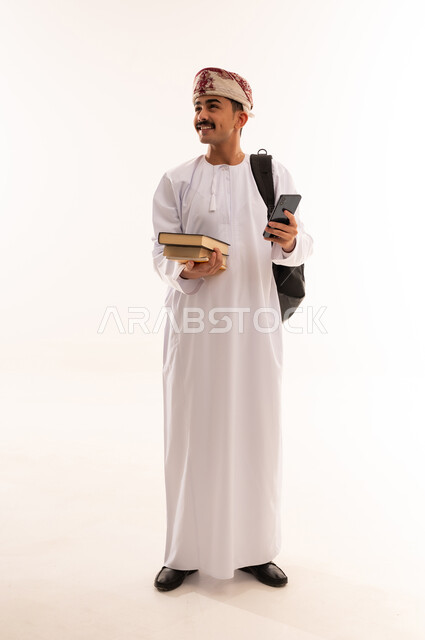 الدردشة والتواصل مع الاصدقاء، الاستفادة من الأجهزة والتكنولوجيا الحديثة في الاطمئنان على الأقارب، بورتريه لرجل عربي خليجي عماني يرتدي الدشداشة والكمه و الحقيبة ويمسك الهاتف المحمول بإيماءات السعادة، الجسم بالطول الكامل، خلفية بيضاء
