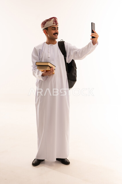 التواصل مع الأهل والأصدقاء وسماع أخبارهم، الاستفادة من الأجهزة والتكنولوجيا الحديثة في الاطمئنان على الأقارب، بورتريه لرجل عربي خليجي عماني يرتدي الدشداشة والكمه و الحقيبة ويمسك الهاتف المحمول و الكتب بإيماءات السعادة، الجسم بالطول الكامل، خلفية بيضاء