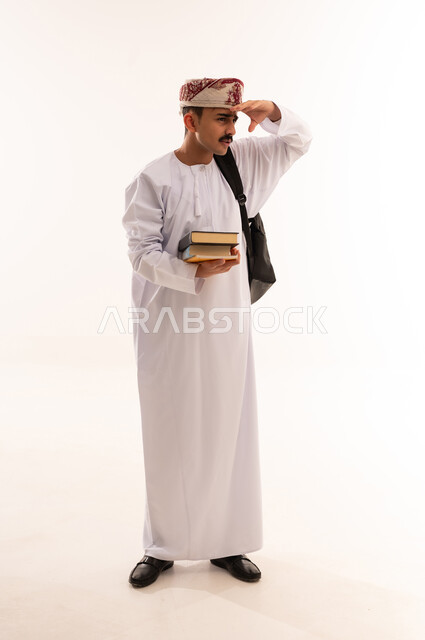 النظر لمكان بعيد, تعابير محاولة رؤية شيء ما, مفهوم التطلع للمستقبل, التعليم الجامعي في عمان, بورتريه لطالب عربي خليجي عماني يرتدي الدشداشة والكمة يحمل حقيبة على ظهره والكتب الجامعية بيده, الجسم بالطول الكامل، خلفية بيضاء