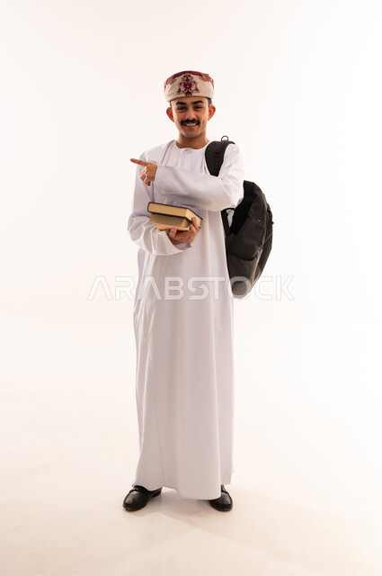التعليم الاكاديمي في سلطنة عمان،الإشارة بإصبع السبابة إلى شيء ما, الاستعداد للعام الجامعي الجديد, بورتريه لطالب جامعي عربي خليجي عماني يرتدي الدشداشة والكمة يحمل حقيبة على ظهره والكتب بيديه, الجسم بالطول الكامل, خلفية بيضاء