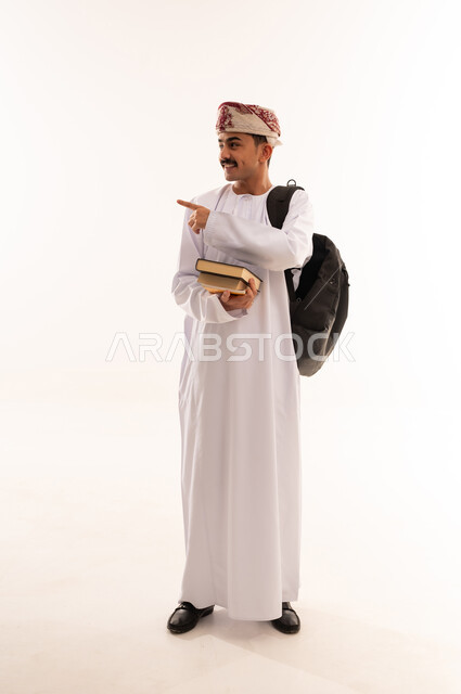 الإشارة بإصبع السبابة إلى شيء ما, الاستعداد للعام الجامعي الجديد, التعليم الاكاديمي في سلطنة عمان, بورتريه لطالب جامعي عربي خليجي عماني يرتدي الدشداشة والكمة ينظر إلى شيء ما يحمل حقيبة على ظهره والكتب بيديه, الجسم بالطول الكامل, خلفية بيضاء