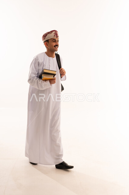الاستعداد للعام الجامعي الجديد، المشي الى الجامعة بايماءات  السعاده، بورتريه لطالب عربي خليجي عماني يرتدي الدشداشة والكمة يحمل حقيبة على ظهره و يمسك الكتب ,التعليم العالي في الجامعات والكليات الأكاديمية في سلطنة عمان، الجسم بالطول الكامل، خلفية بيضاء