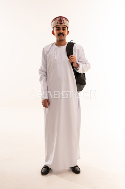 الاستعداد للعام الجامعي الجديد، النظر بإيماءات السعادة والسرور,التعليم العالي في الجامعات والكليات الأكاديمية في سلطنة عمان, بورتريه لطالب عربي خليجي عماني يرتدي الدشداشة والكمة يحمل حقيبة على ظهره وبيديه الكتب, الجسم بالطول الكامل، خلفية بيضاء
