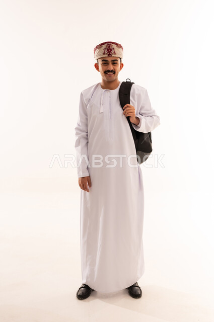النظر بإيماءات السعادة والسرور, الاستعداد للعام الجامعي الجديد, التعليم العالي في الجامعات والكليات الأكاديمية في سلطنة عمان, بورتريه لطالب عربي خليجي عماني يرتدي الدشداشة والكمة يحمل حقيبة على ظهره وبيديه الكتب, الجسم بالطول الكامل، خلفية بيضاء
