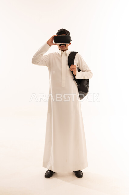Watching 3D movies through modern virtual reality glasses, interaction, fun and entertainment by watching movies, portrait of a Saudi Arabian Gulf student wearing traditional clothing and a bag using virtual world glasses, full body length, white background
