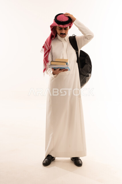 الاستعداد للذهاب الى الجامعة، بورتريه لطالب جامعي عربي خليجي سعودي مبتسم يرتدي الزي التقليدي والحقيبة يحمل بيده الكتب الجامعية يقوم بالتفكير في شيء ما، التعليم الجامعي في المملكة العربية السعودية ، الجسم بالطول الكامل، خلفية بيضاء