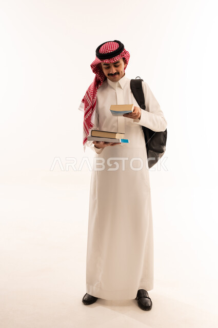 متابعة المحاضرات الجامعية، بورتريه لطالب جامعي عربي خليجي سعودي مبتسم يرتدي الزي التقليدي والحقيبة الجامعية ويحمل بيده الكتب الجامعية ، الاستعداد للذهاب الى الجامعة، التعليم الجامعي في المملكة العربية السعودية ، الجسم بالطول الكامل، خلفية بيضاء