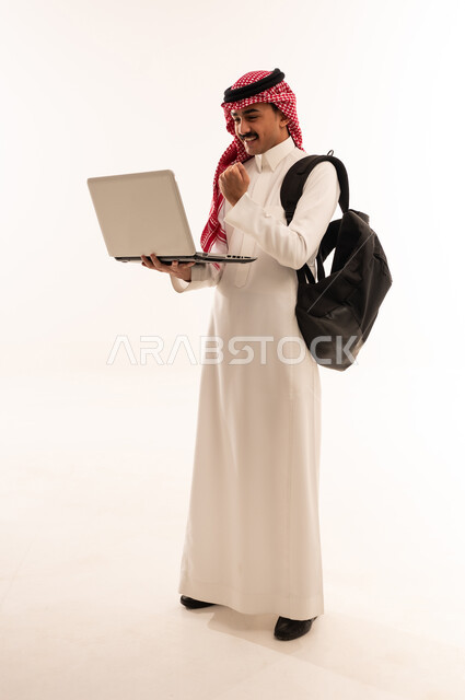 البحث والمطالعة وتصفح التطبيقات الالكترونية، متابعة الدرجات من خلال الحاسب المحمول، بورتريه لطالب جامعى عربي خليجي سعودي يحمل حقيبة الظهر، الاستعداد للعام الدراسي الجامعي الجديد، ايماءات الوجه واليد تدل على النجاح والامتياز، الجسم بالطول الكامل، خلفية بيضاء