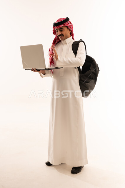 مفهوم التعليم عن بعد، استخدام الأجهزة التقنية الحديثة لمتابعة المحاضرات الجامعية, بورتريه لطالب جامعي عربي خليجي سعودي مبتسم يرتدي الزي التقليدي والحقيبه الجامعية يحمل بيده الحاسوب المحمول و يقوم بمكالمه فيديو, الجسم بالطول الكامل، خلفية بيضاء