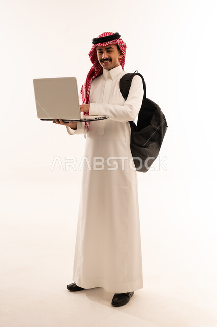 مفهوم التعليم عن بعد، استخدام الأجهزة التقنية الحديثة لمتابعة المحاضرات الجامعية، المناهج و المواد التعليمية في السعودية, بورتريه لطالب جامعي عربي خليجي سعودي مبتسم يرتدي الزي التقليدي و الحقيبه الجامعية يحمل بيده الحاسوب المحمول, الجسم بالطول الكامل، خلفية بيضاء
