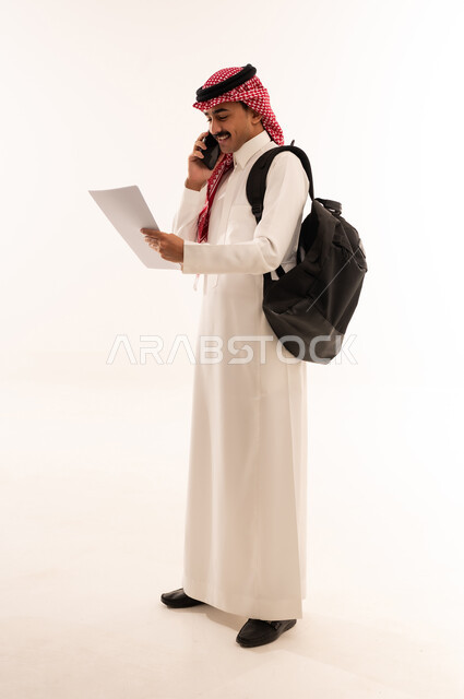 استخدام الأجهزة التقنية الحديثة, إجراء اتصال هاتفي عبر الجوال, التعليم الجامعي في السعودية, بورتريه لطالب جامعي عربي خليجي سعودي يرتدي الزي التقليدي يحمل حقيبة على ظهره ويمسك بيديه ورق جامعي والهاتف المحمول, الجسم بالطول الكامل, خلفية بيضاء