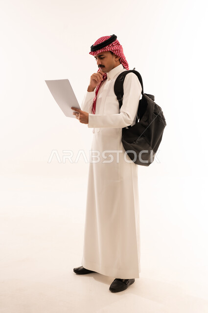 التعليم في المملكة العربية السعودية ، إيماءات وجه ويدين تدل على التفكير والحيرة ، بورتريه لطالب جامعي عربي يرتدي الزي الخليجي السعودي و يحمل حقيبه يمسك الاوراق الدراسية في يده ، الإستعداد للمحاضرات الجامعية ،الجسم بالطول الكامل، خلفية بيضاء