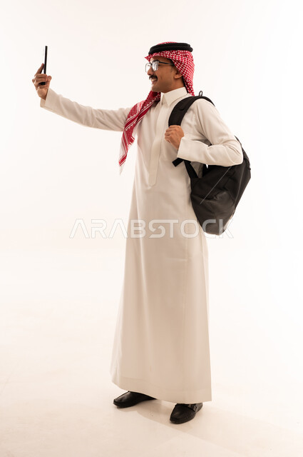 الاستمتاع في التحدث مع الاصدقاء، ايماءات السعادة والسرور، استخدام التقنيات الحديثة والتكنولوجيا، بورتريه لطالب عربي خليجي جامعي يرتدي الثوب التقليدي يحمل حقيبه على ظهره و يقوم بالتواصل مع الاهل والاقارب عبر الهاتف المحمول، الجسم بالطول الكامل، خلفية بيضاء